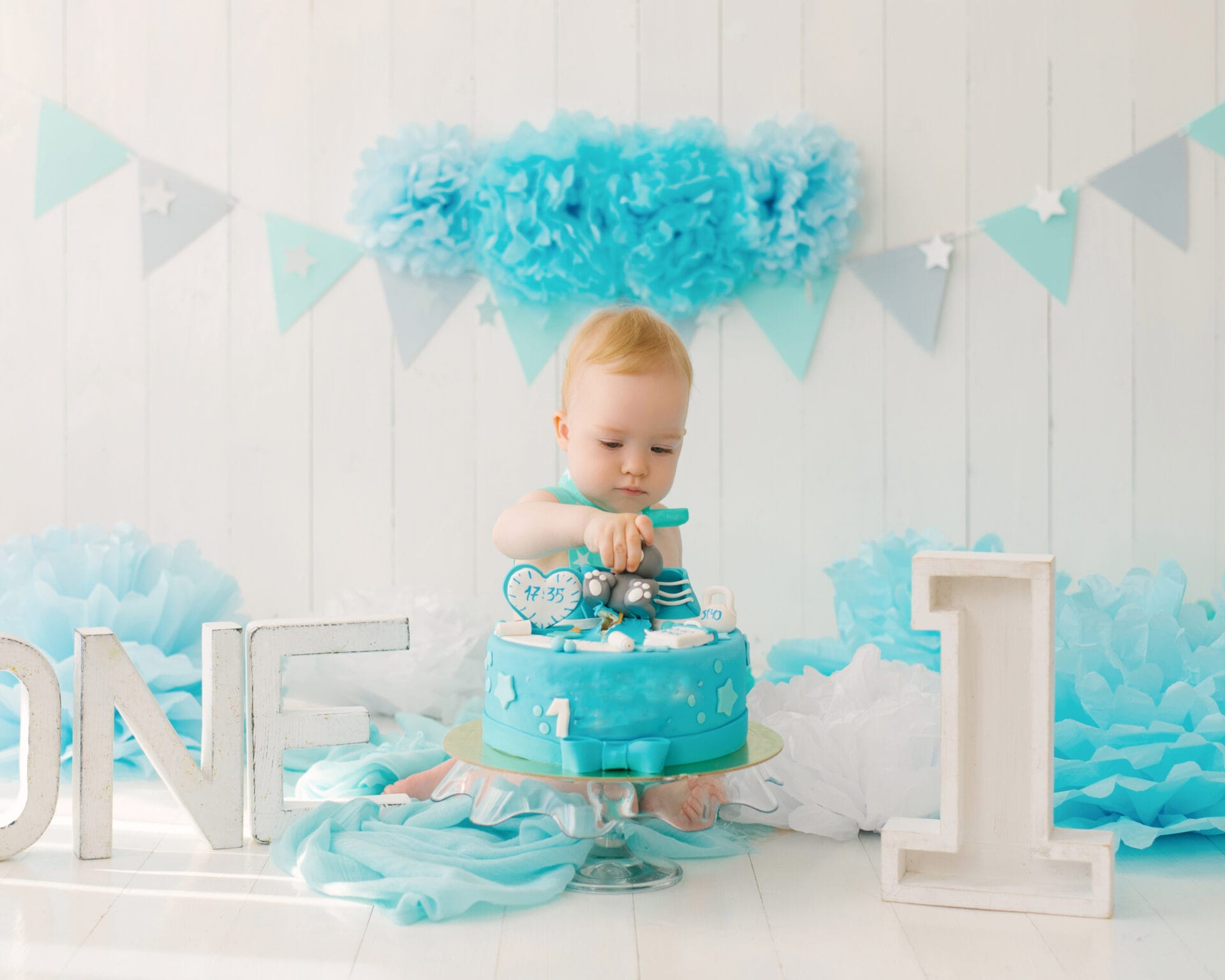 baby dressed in blue clothing picking at a birthday cake to celebrate their first birthday