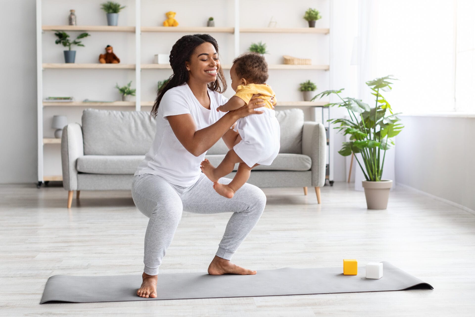 Workout With Baby. Cheerful Black Woman Exercising At Home With Infant Son, Sporty African American Female Training On Fitness Mat, Holding Her Cute Toddler Child And Making Squats